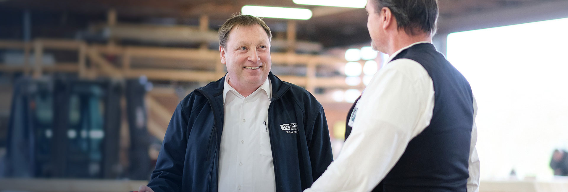 Volker Rozek im Gespräch mit einem Kunden in der Werkshalle über mögliche Prozessoptimierungen im mittelständischen Produktionsbetrieb. Im Hintergrund sind Lagerflächen und Maschinen sichtbar.